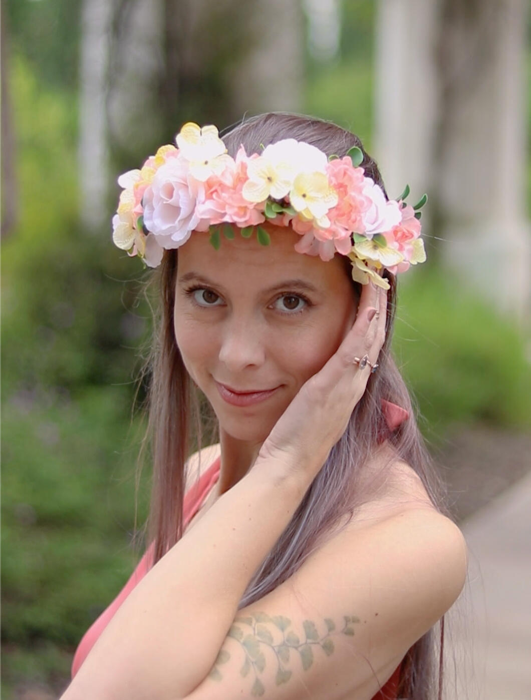 A picture of a woman in her 30's wearing a peach and yellow flower crown smiling at the camera with one hand touching the crown along the side of her face.